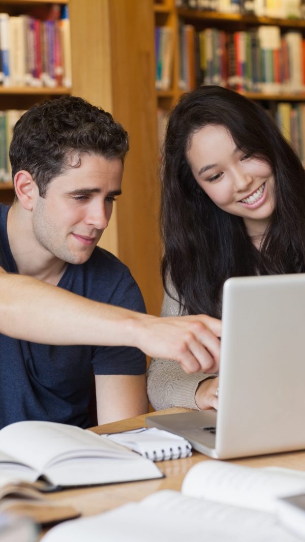 Two students sitting at a desk in a library while having fun and using the laptop